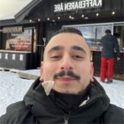 Person i vinterkl&auml;der som tar en selfie framf&ouml;r en restaurang i sn&ouml;n, med en annan person i bakgrunden.
