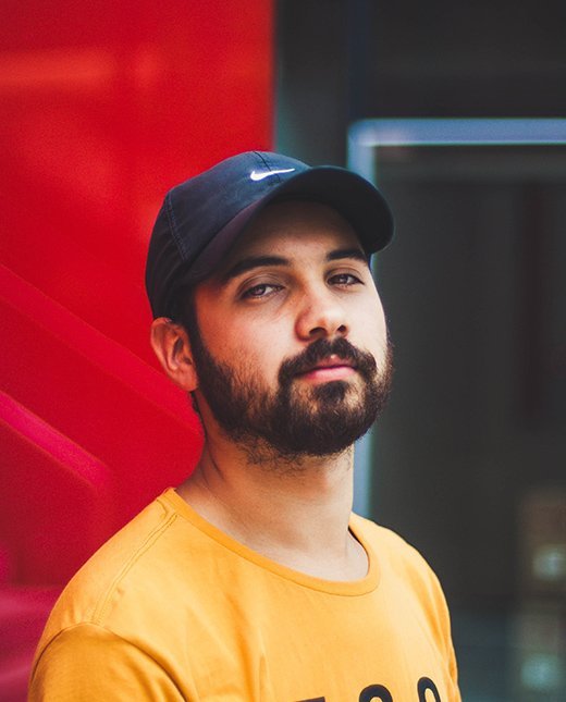 Man med sk&auml;gg och keps, kl&auml;dd i orange tr&ouml;ja, poserar framf&ouml;r en r&ouml;d bakgrund, relaterat till webdesign och kundrecensioner p&aring; Enkelwebb.se.