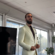 Man i ljus kostym som st&aring;r i ett kontorsutrymme med f&ouml;nster och en stor TV-sk&auml;rm i bakgrunden, representerar professionell webbutveckling och kreativitet.