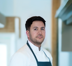 Kock i vit uniform och f&ouml;rkl&auml;de, st&aring;ende i modern k&ouml;ksmilj&ouml;, vilket speglar Enkelwebbs fokus p&aring; professionella och anv&auml;ndarv&auml;nliga l&ouml;sningar f&ouml;r f&ouml;retag.